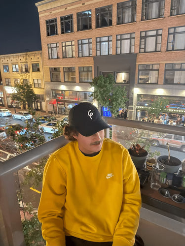 Person wearing a yellow sweatshirt and black 6th Panel Staple hat sitting on a balcony with cityscape view.