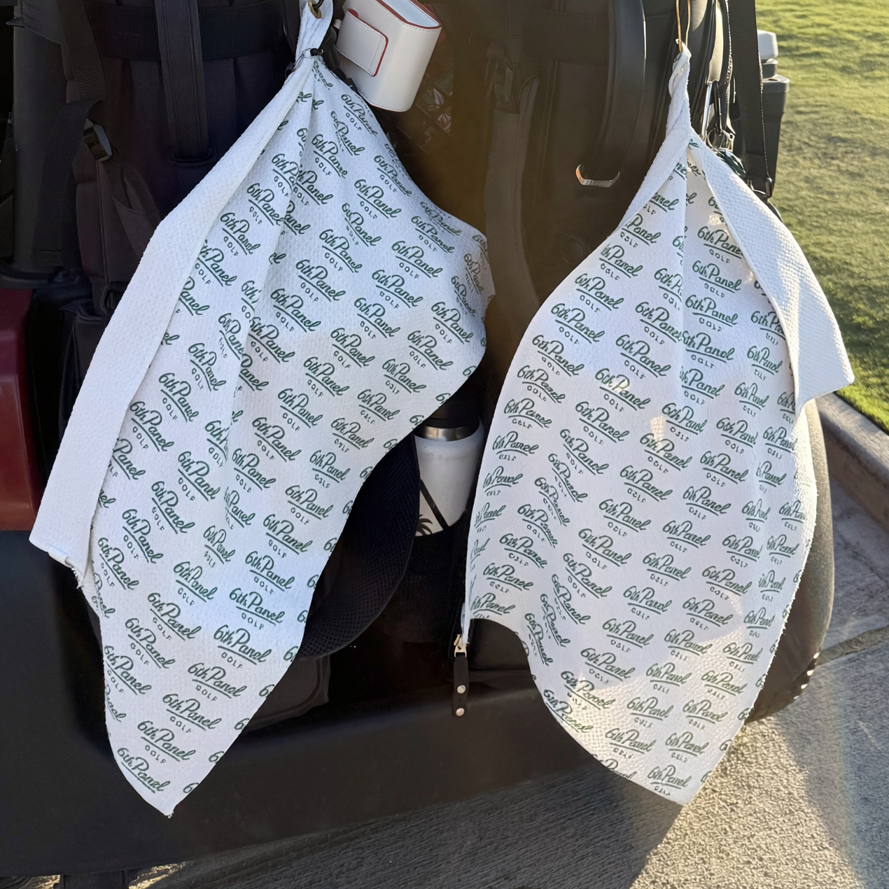 Two white golf bibs with a repeating dark green '6th Panel' logo pattern, hanging from the open rear of a black golf cart outdoors.