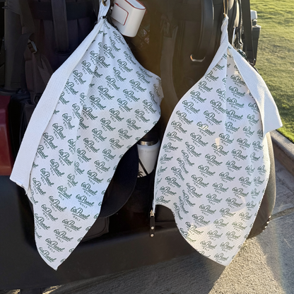 Two white golf bibs with a repeating dark green '6th Panel' logo pattern, hanging from the open rear of a black golf cart outdoors.